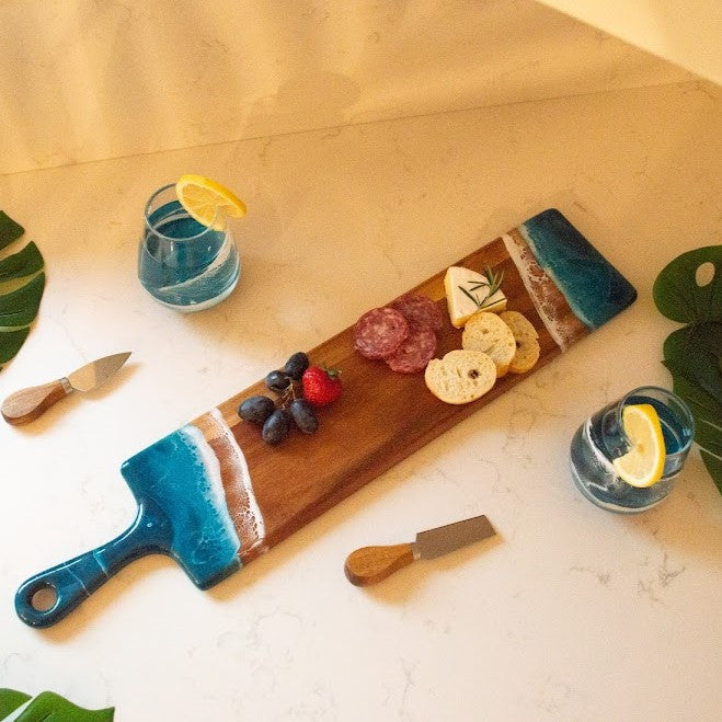 Wooden serving board with blue handle, sliced fruits, and drinks on a marble surface.