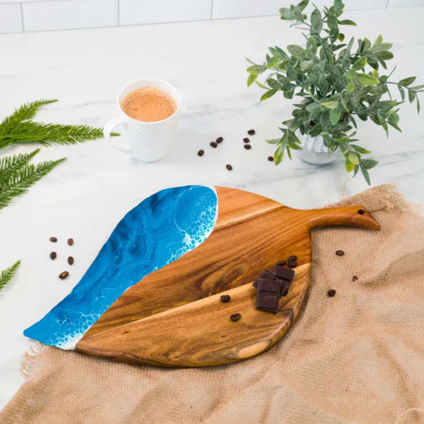 Wooden cutting board with blue resin design on a white surface with coffee beans and a plant.