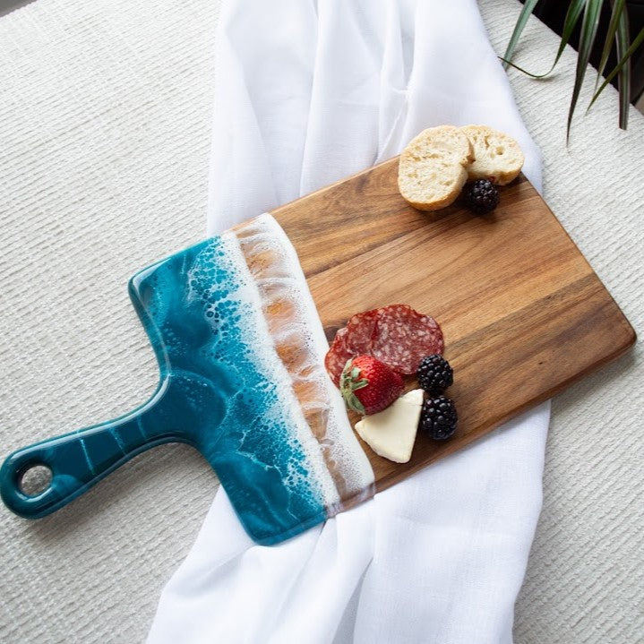 Wooden cutting board with a blue handle, sliced bread, cheese, and fruit on a white cloth.