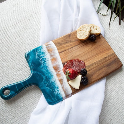 Wooden cutting board with a blue handle, sliced bread, cheese, and fruit on a white cloth.