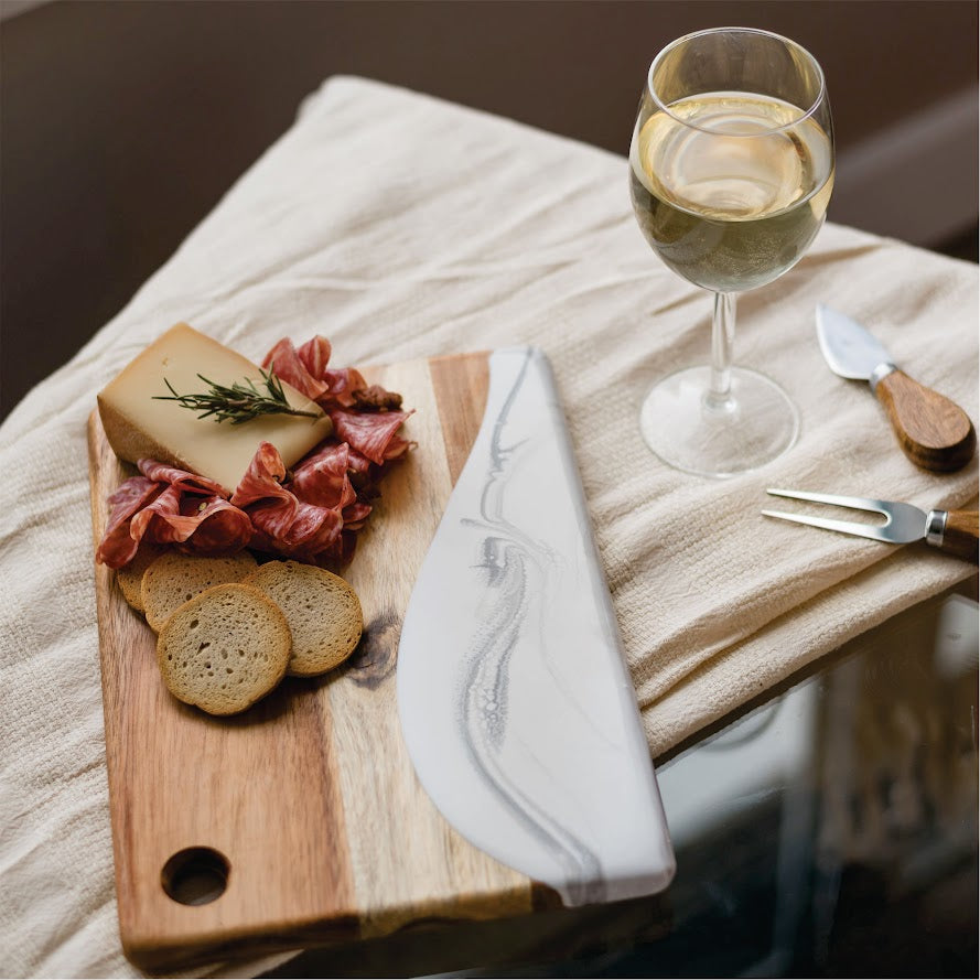 Wooden cutting board with cured meats, bread, and a glass of white wine on a soft surface.