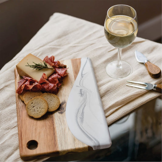 Wooden cutting board with cured meats, bread, and a glass of white wine on a soft surface.