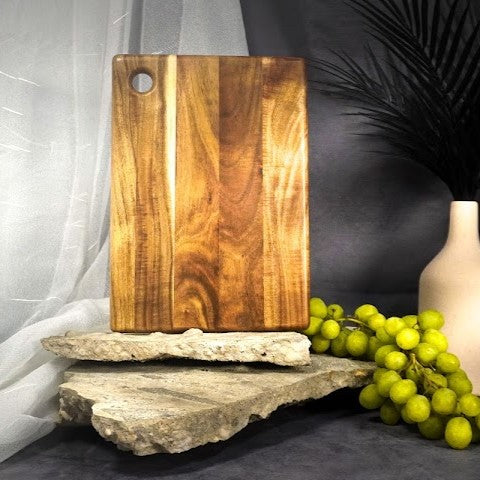 Wooden cutting board on a stone surface with grapes and a vase in the background